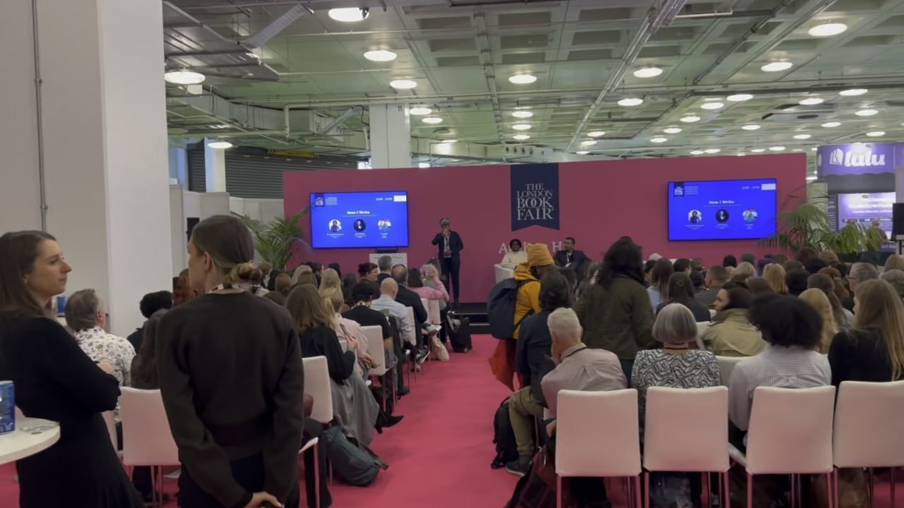 A crowded seminar at the London Book Fair, with a speaker on a pink stage and screens either side